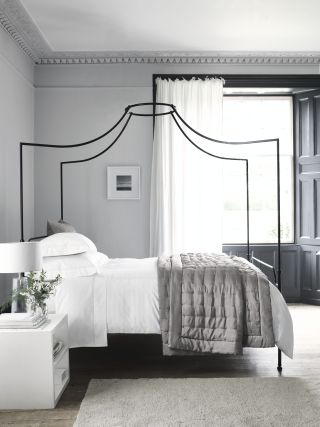 Bedroom by The White Company, pale gray walls, white bedside, four poster metal bed, gray quilt, white bedding, wooden floorboards, gray rug