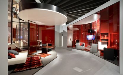 View of Lazzarini Pickering’s room installation featuring red walls, seating in different colours, a rug, a tv and a table. There is a light grey pathway running through the room