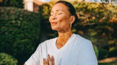 A woman stands outside with her eyes closed and hands clasped together in prayer pose. 