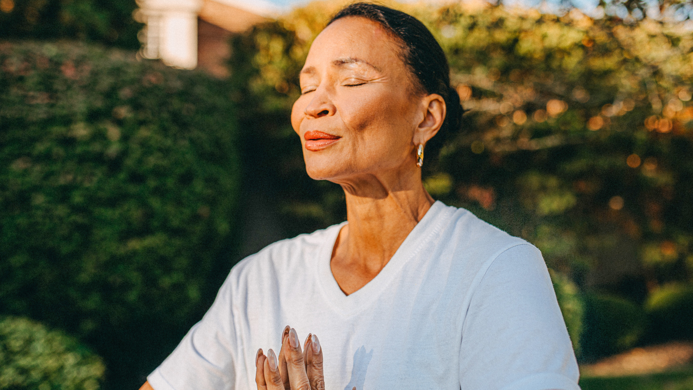 A woman stands outside with her eyes closed and hands clasped together in prayer pose. 
