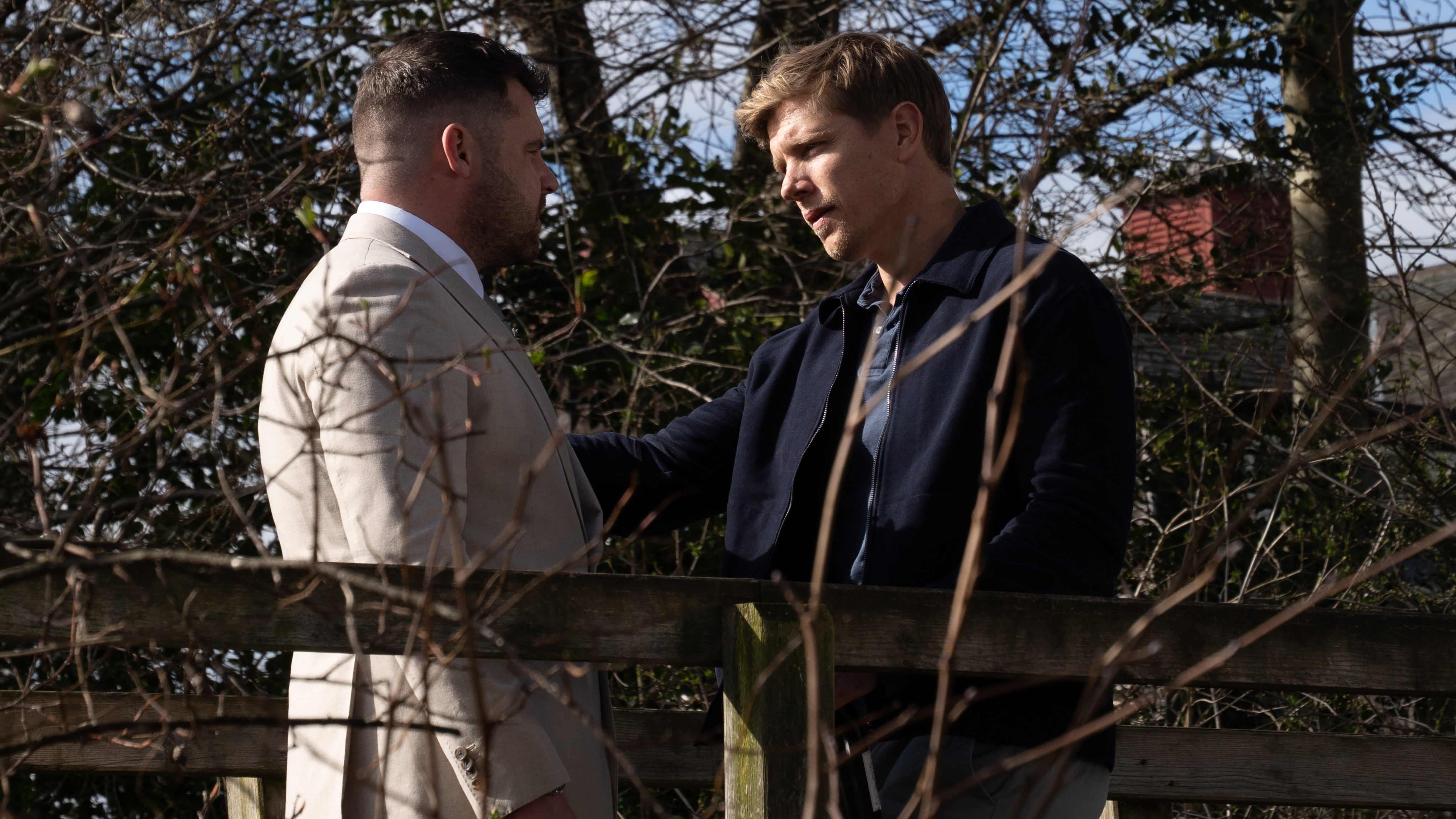 Aaron Dingle and Robert Sugden standing together on a bridge. Aaron is wearing a cream wedding suit.