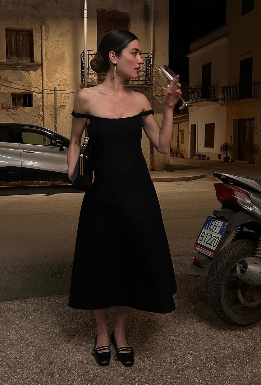 A woman standing outside with a wine glass, wearing a black off-the-shoulder midi dress with black kitten heels, a black clutch, and silver earrings
