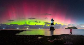 vibrant ribbons of green and pink auroras spread across the sky with a lighthouse in the center and a person to the right with their arms in the air.