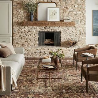 A farmhouse-style living room with a stone fireplace, maroon area rug, and a wooden shelf