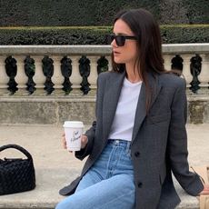 French woman sitting on bench wearing white shirt, jeans, and grey blazer, holding coffee cup and wearing sunglasses.