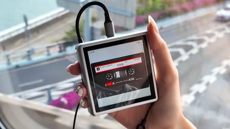 Hand with long nails holding Anbernic RG Rotate with USB cable coming out of top and cassette tape animation on screen.