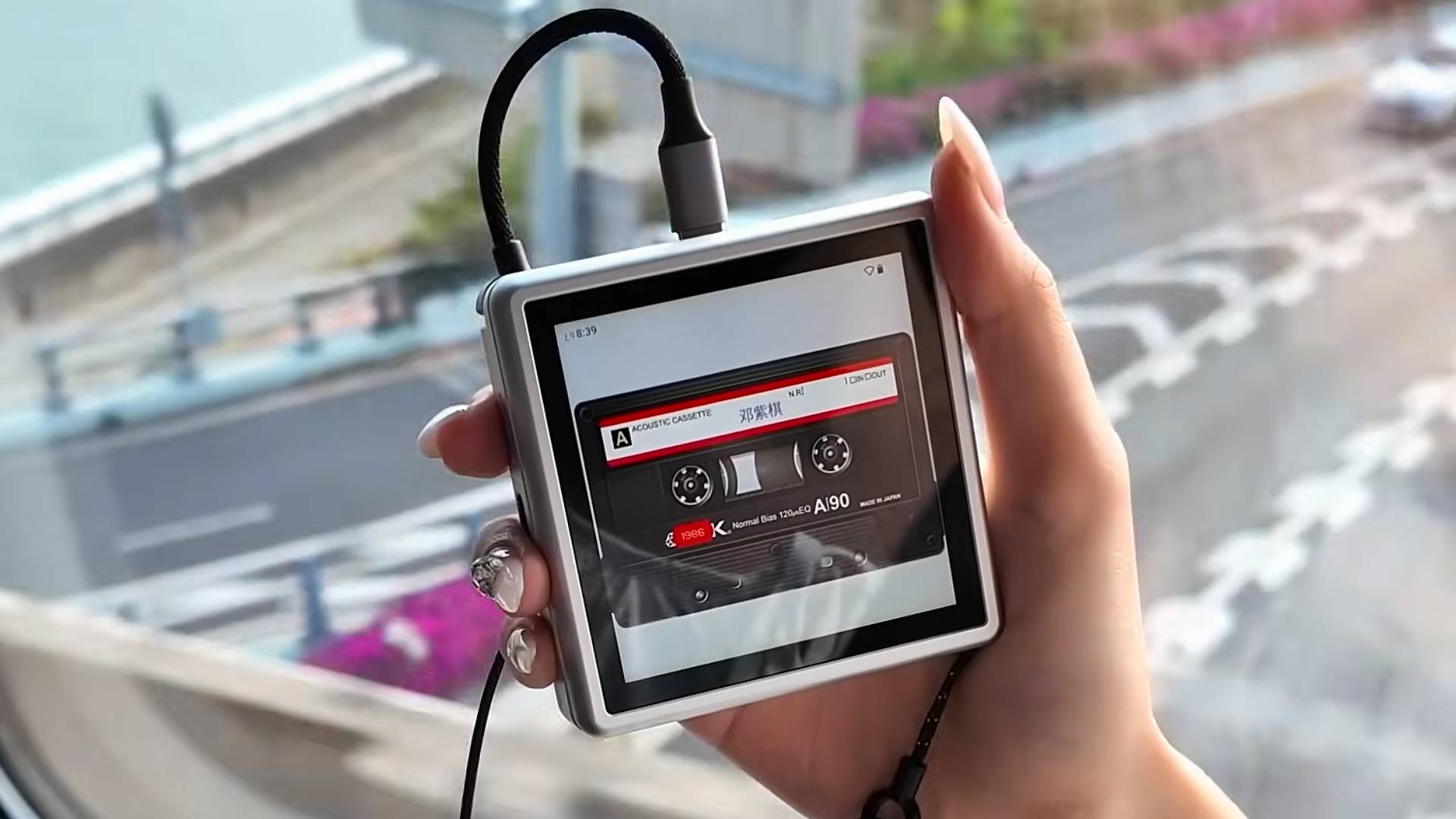 Hand with long nails holding Anbernic RG Rotate with USB cable coming out of top and cassette tape animation on screen.