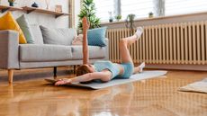 A woman lies on her back performing a dead bug exercise on an exercise mat in her living room. Her left leg and right arm are extended along the floor, while her left arm and right leg are elevated.