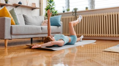 A woman lies on her back performing a dead bug exercise on an exercise mat in her living room. Her left leg and right arm are extended along the floor, while her left arm and right leg are elevated.
