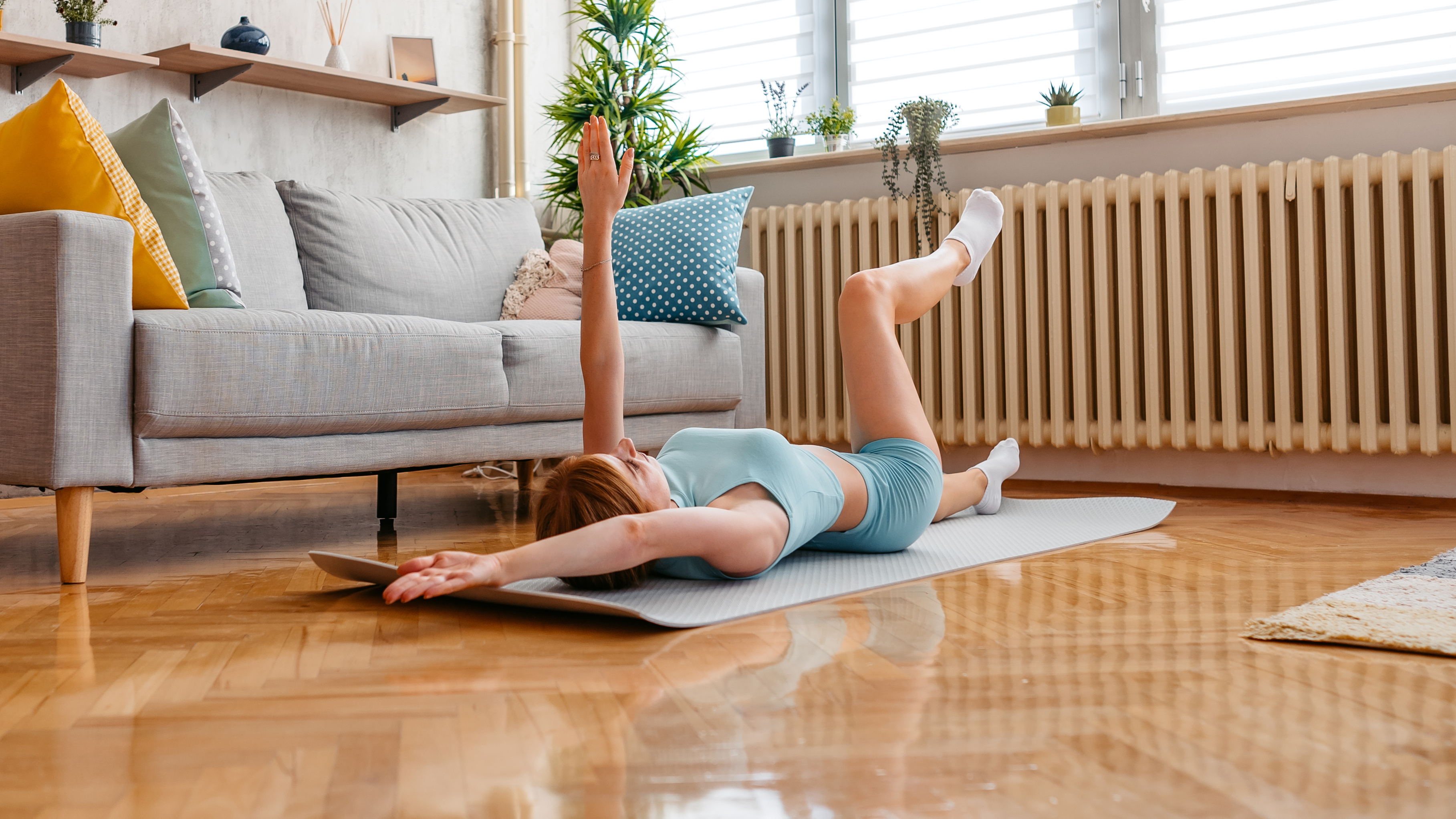 A woman lies on her back performing a dead bug exercise on an exercise mat in her living room. Her left leg and right arm are extended along the floor, while her left arm and right leg are elevated.