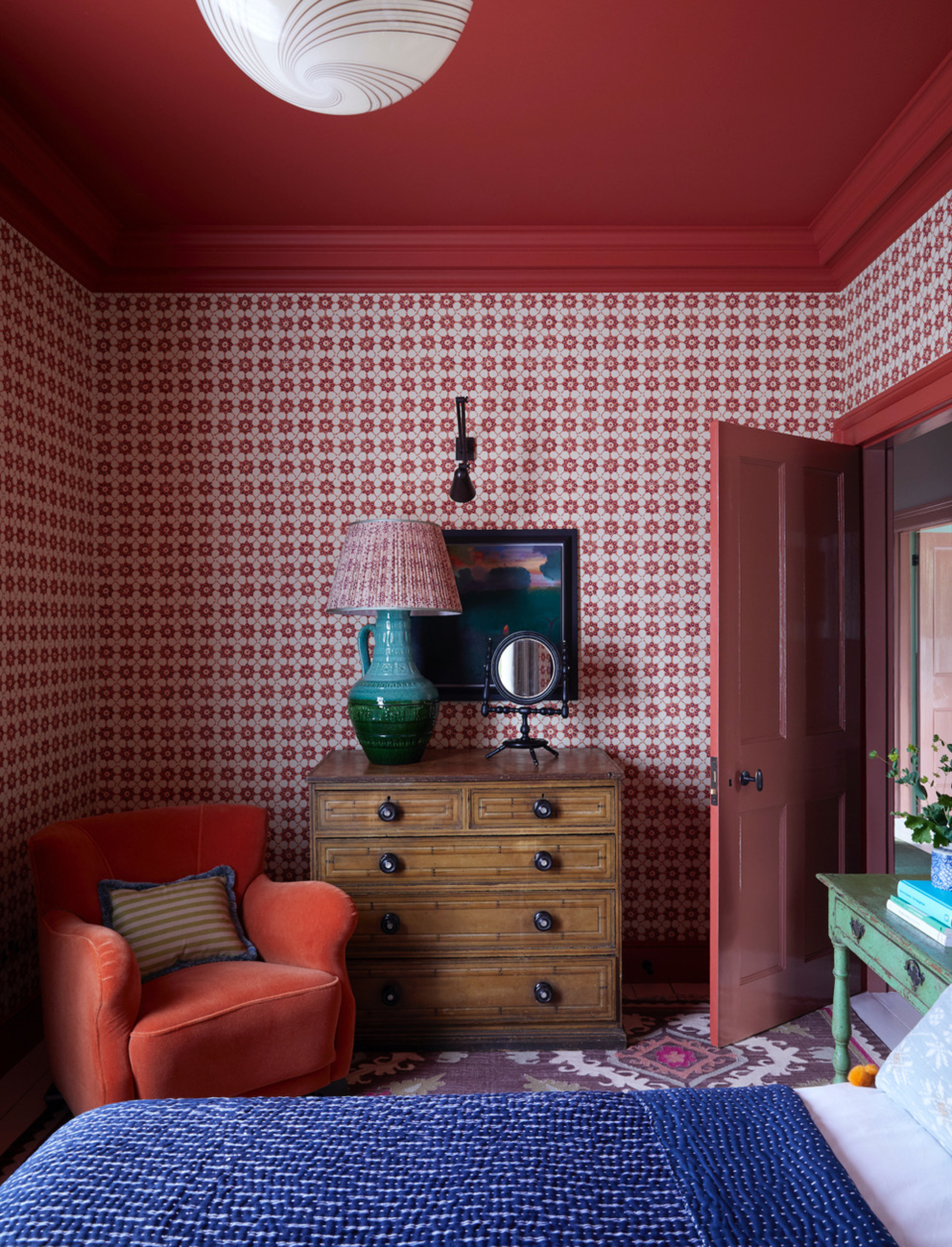 bedroom with red ceiling, red patterned wallpaper, blue bedcover and white sheets, patterned carpet, red armchair with striped cushion, and timber chest of drawers with mirror and lamp styled on top with artwork hung on the wall above