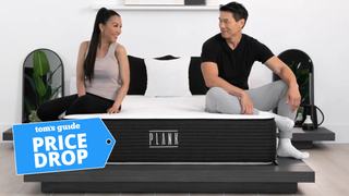 The image shows a man and woman sitting on the Plank Firm Luxe mattress in a sleek, white bedroom