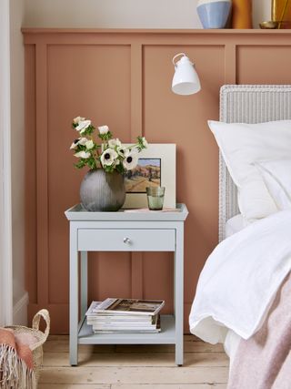 terracotta painted wall panelling in bedroom, pale grey nightstand, white wall light, white painted rattan bed, white bedding, pink blanket, shelf on top of paneling, wooden floor
