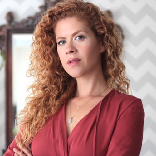 Shula is a white woman with long curly ginger hair. She's pictured wearing a clay red blouse with her arms folded.
