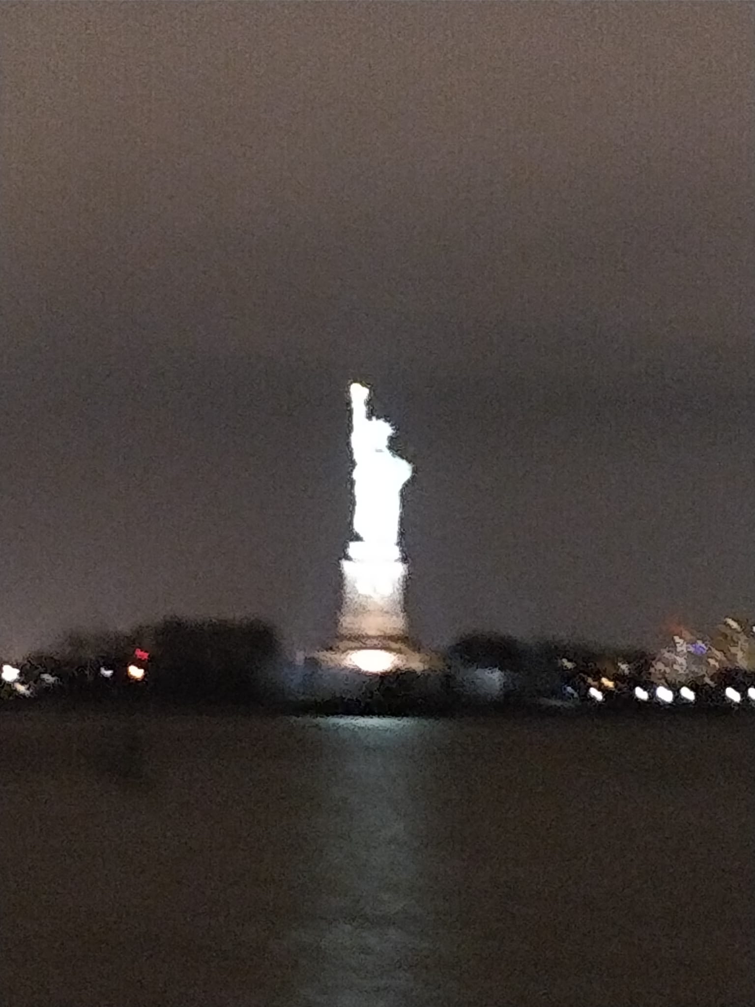 A blurred, overexposed Statue of Liberty glows white against a dark harbor skyline at night.