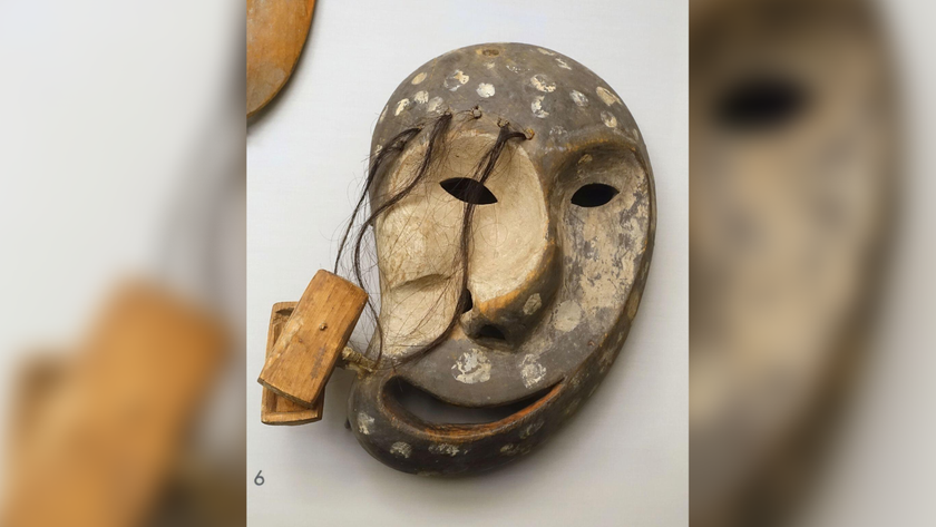 Carved driftwood mask in the shape of a human face but distorted. There is hair coming out of the top of the right eye.