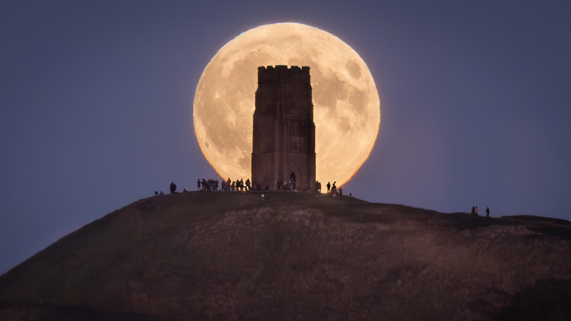 A full moon is pictured rising behind a solitary tower on a hill, around which people have gathered.