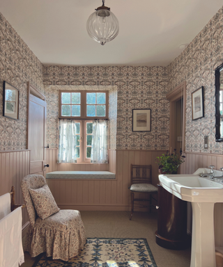 Bathroom with light pink patterned wallpaper and covered skirted chair with rug and wall panelling also in pink and antique wooden chair and white sink and glass pendant ceiling light