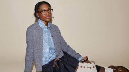 Demi Singleton poses for photo wearing gray cardigan, checkered button-down shirt, navy midi skirt, brown loafers, and a white bucket bag. 