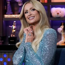 Paris Hilton wearing a blue sparkly dress clasping her hands and smiling with a bookshelf behind her