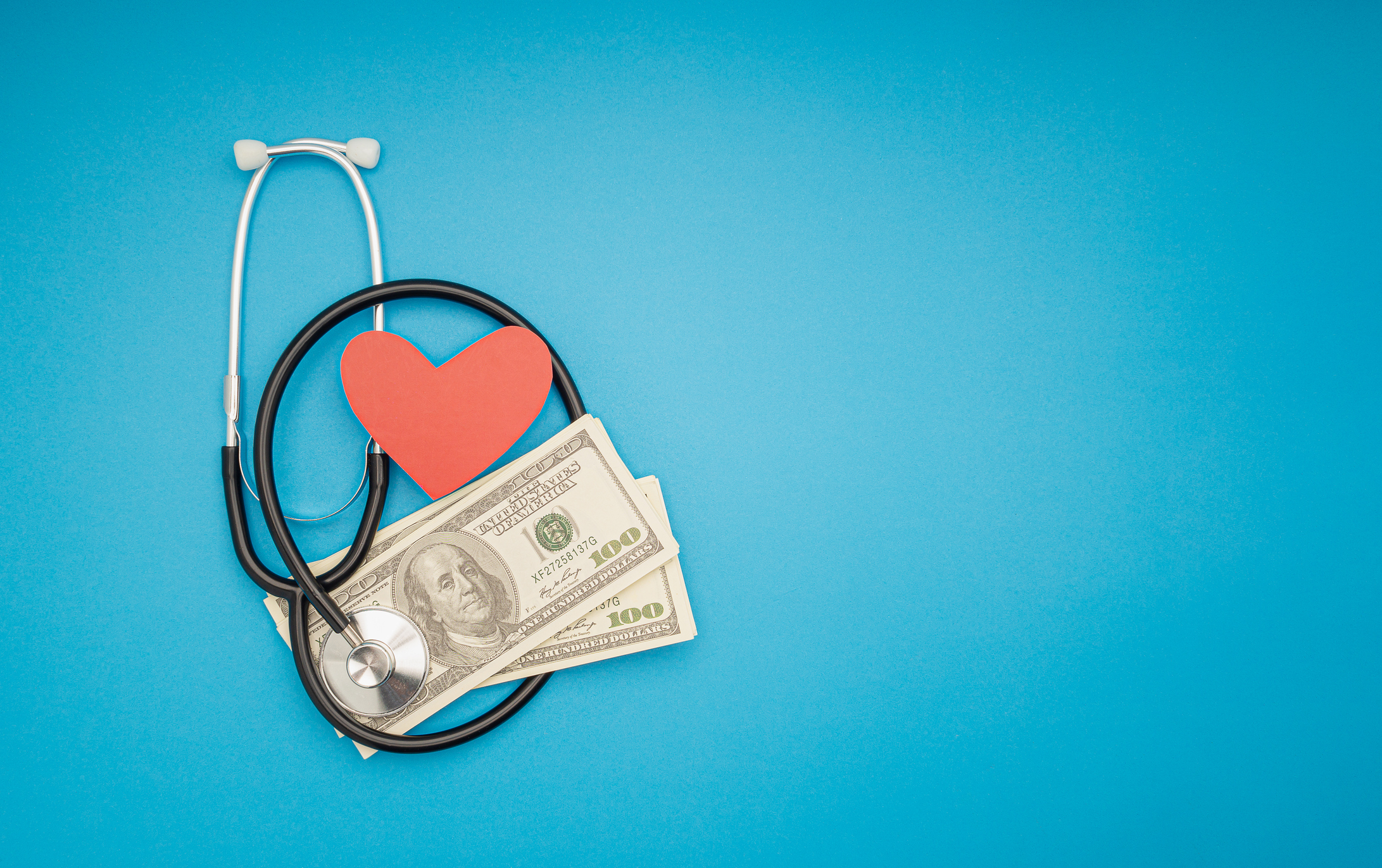 Top view of a stethoscope, US dollar banknotes, and a red heart symbol on a blue background. Space for text. Medical and healthcare concept