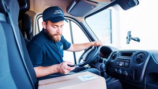 A delivery driver stationary in his van looking at his smartphone with a parcel on the passenger seat