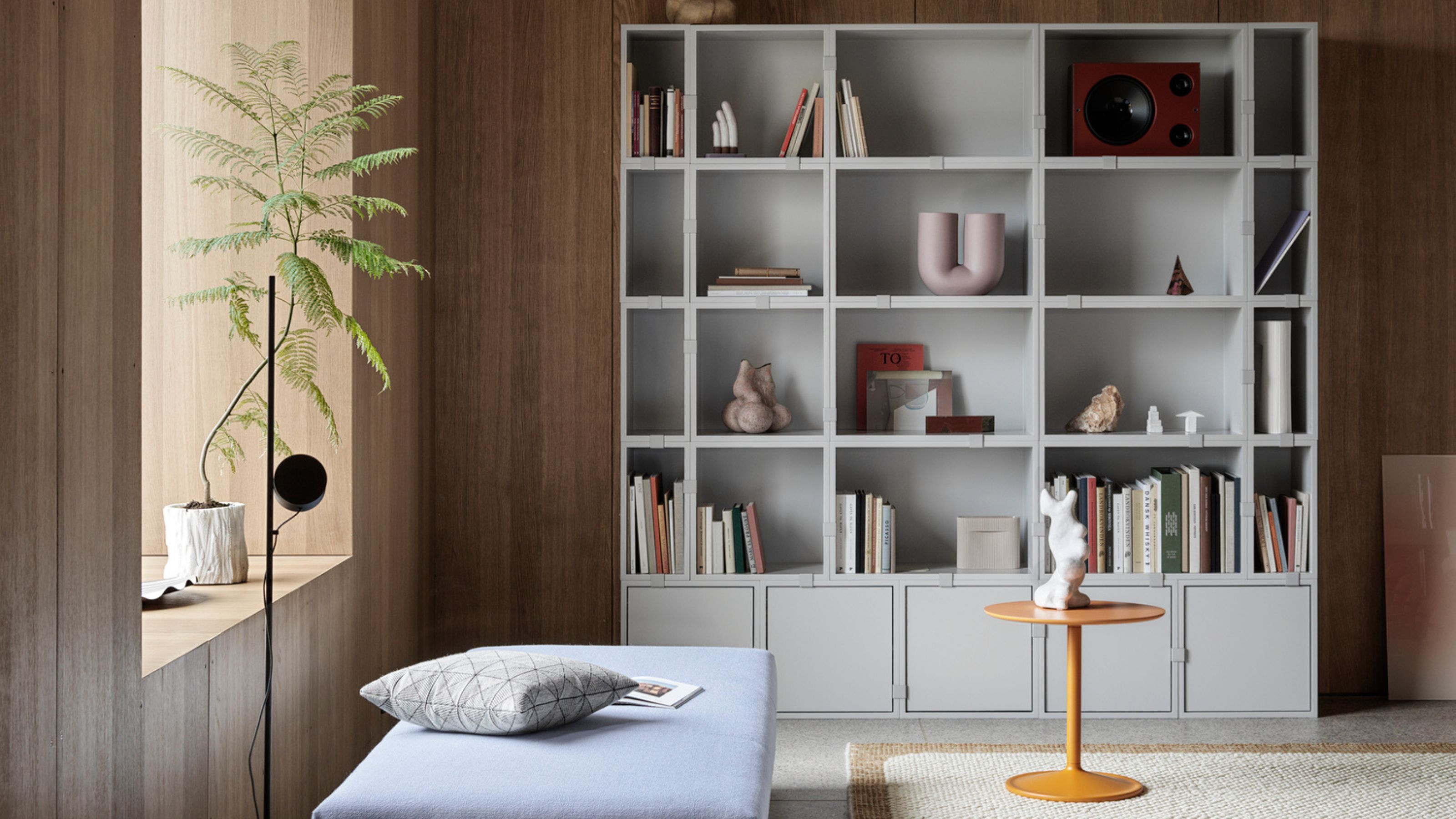 Muuto's shelving system in a modern living room with timber wall panelling, a day bed, white rug, yellow side table, and pot plant on the windowsill