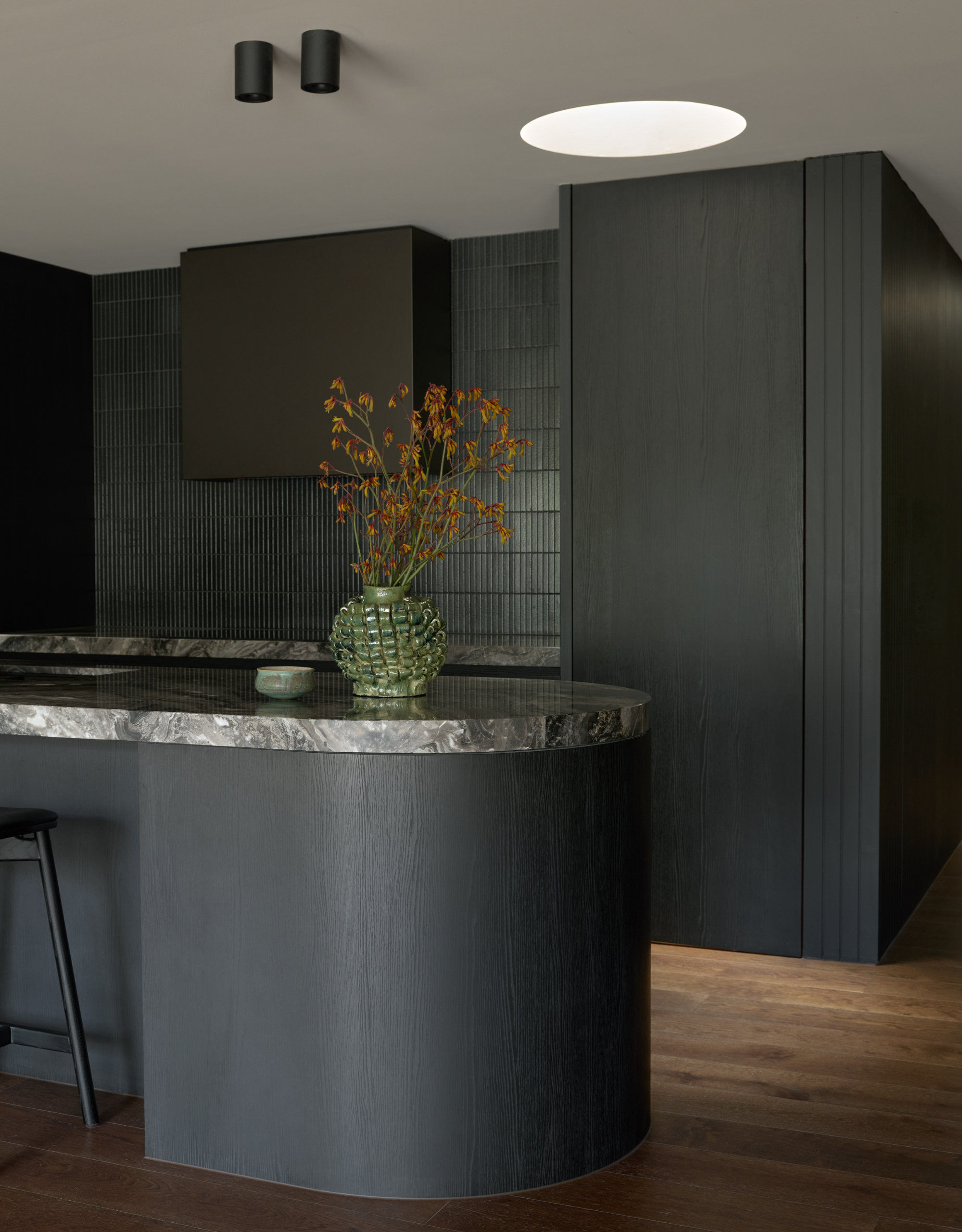 A kitchen with dark wood cabinets, a tiled backsplash, a curved island with a stone top, a green vase, and some stems