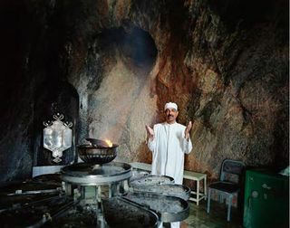 A man dressed in simple white robes with hands raised upwards, stands behind a fire-temple inside a grotto shrine.