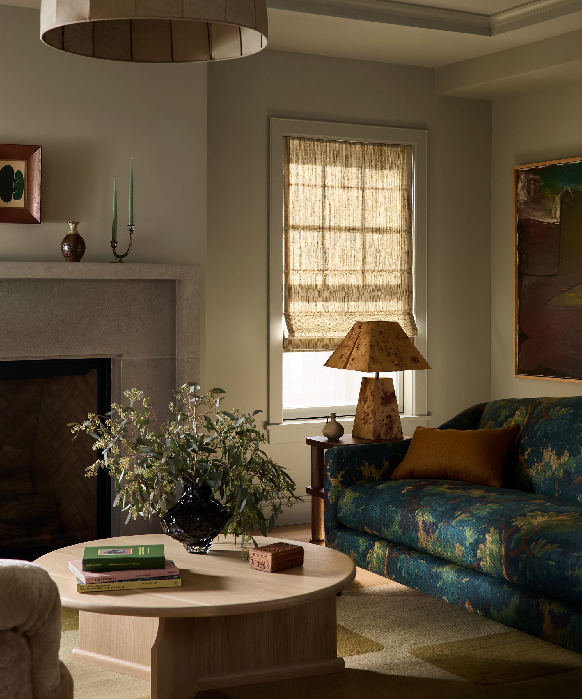 a muted grey green sitting room with a fireplace, small window, tapestry print couch, rounded stone coffee table and a side table with a burl wood lamp