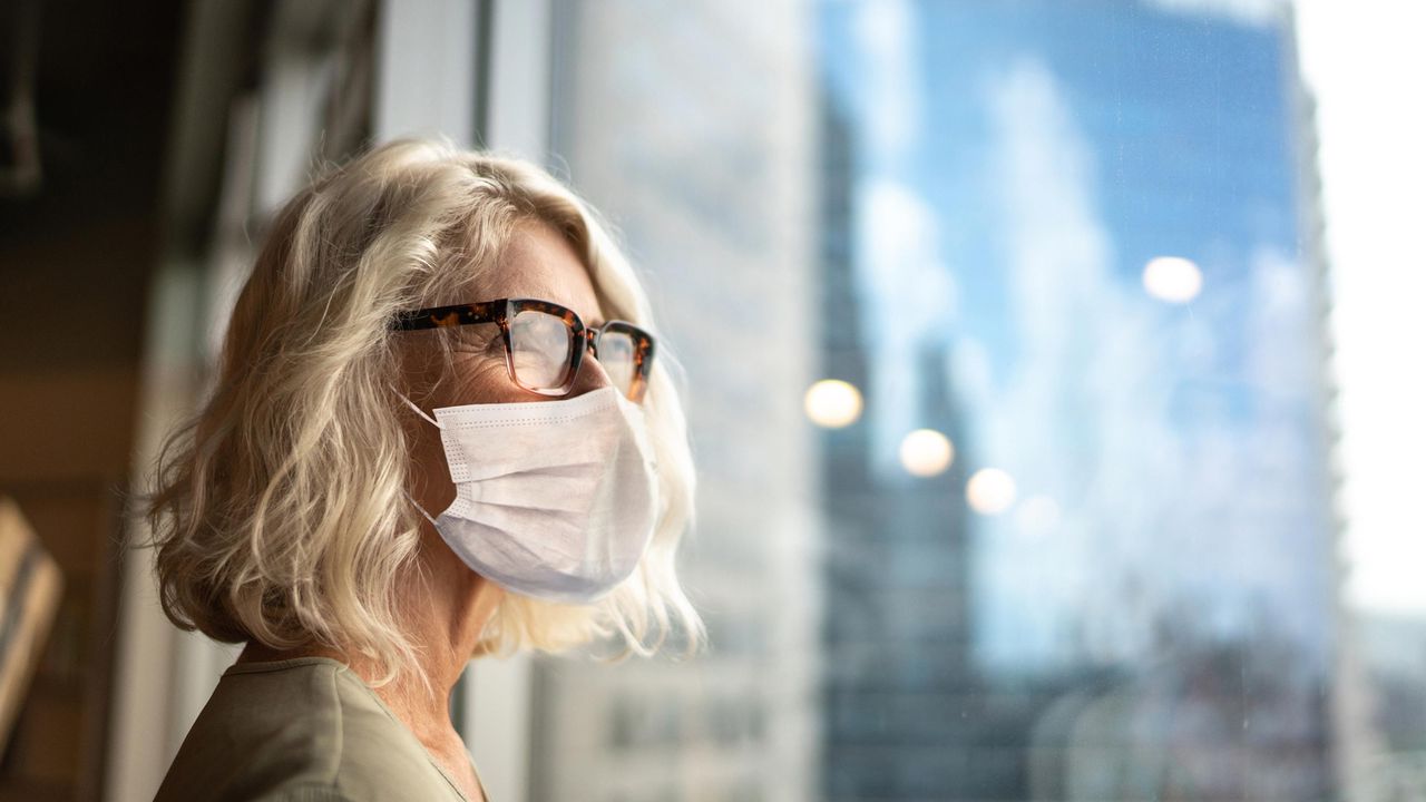 A woman in a business suit wearing a mask looks out an office window.