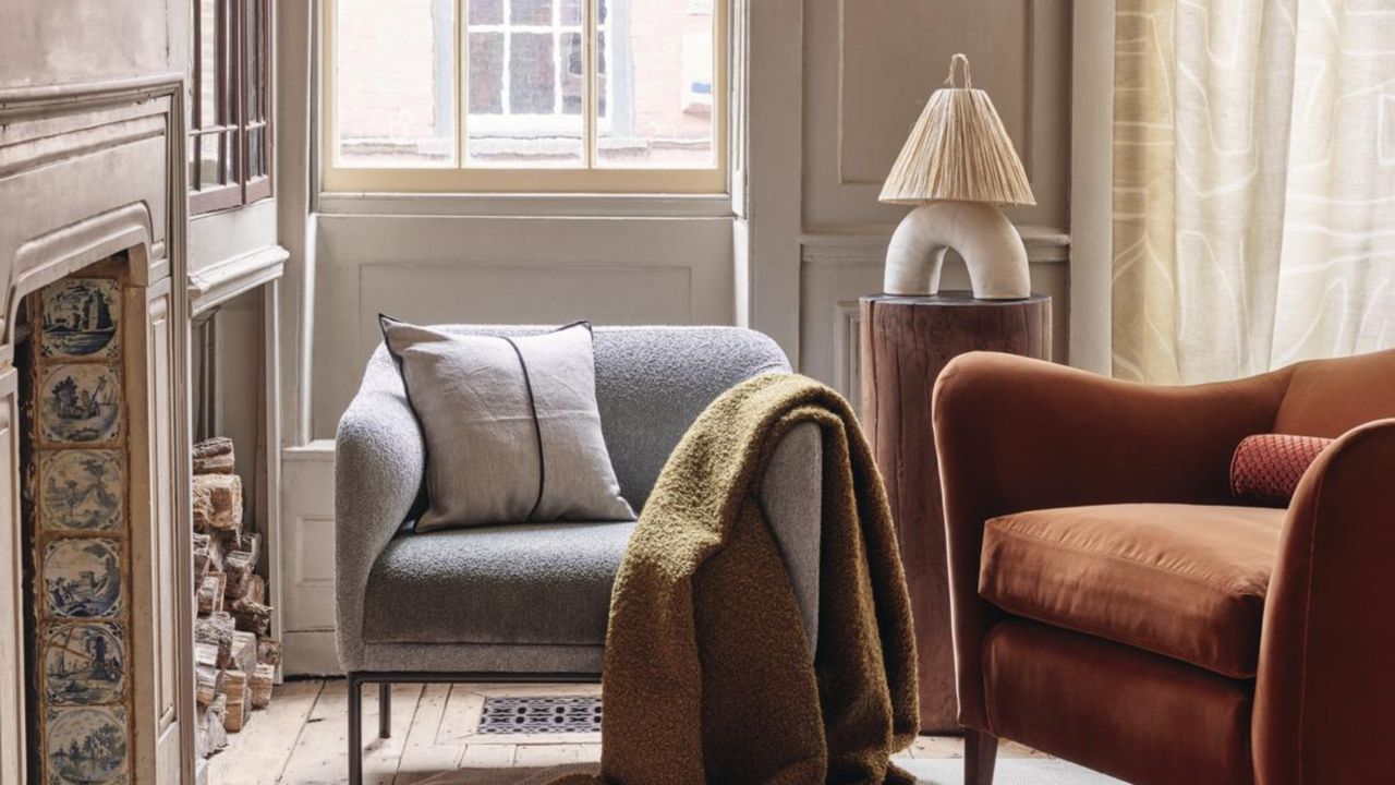 A close up image of a gray accent chair and an orange accent chair positioned facing a fire place. They are in a gray living room and there are a couple windows in view