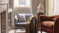 A close up image of a gray accent chair and an orange accent chair positioned facing a fire place. They are in a gray living room and there are a couple windows in view