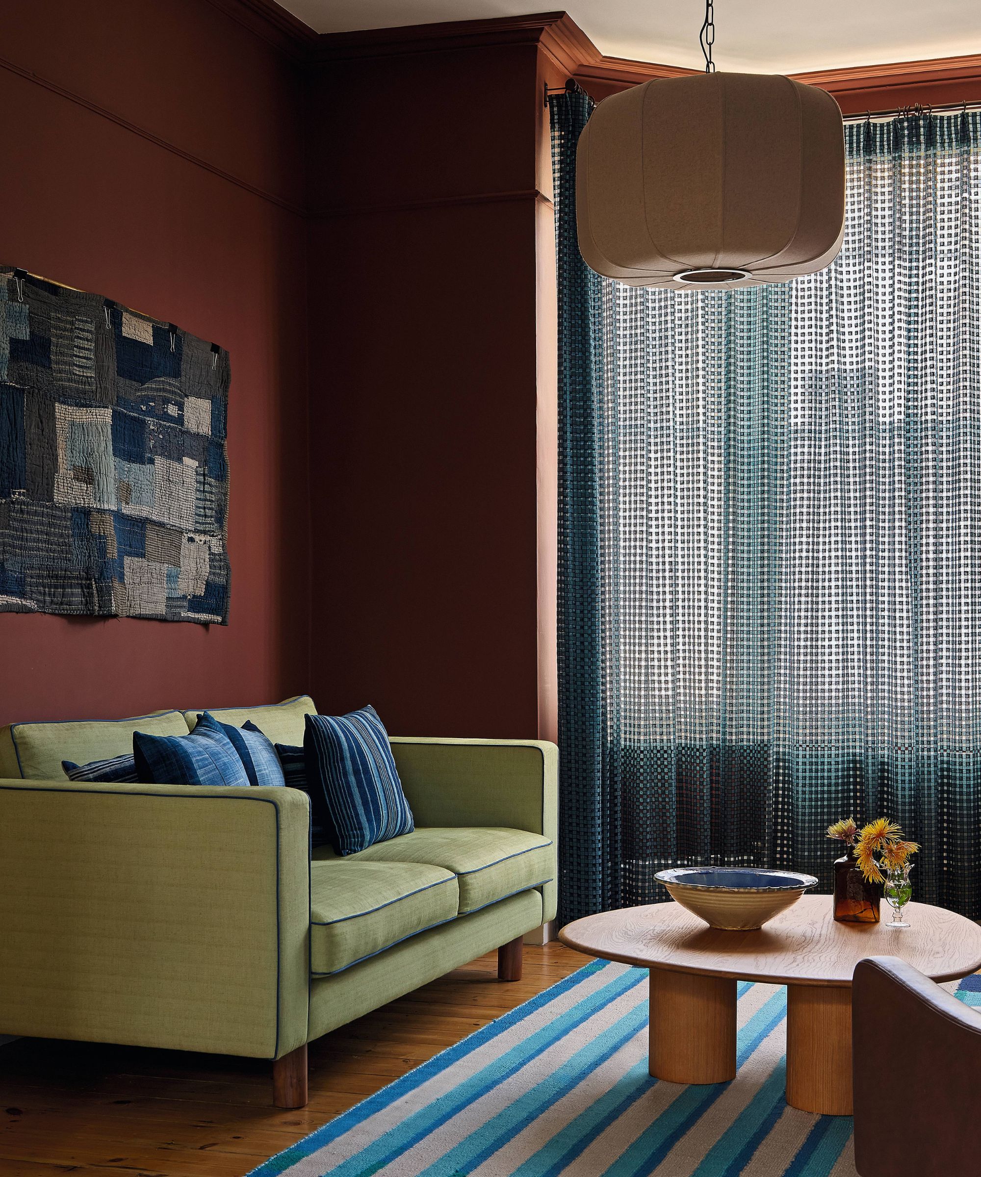 a red painted victorian living room with high ceilings and a bay window. a lime green couch is styled with a blue striped rug and blue sheer drapes at the window
