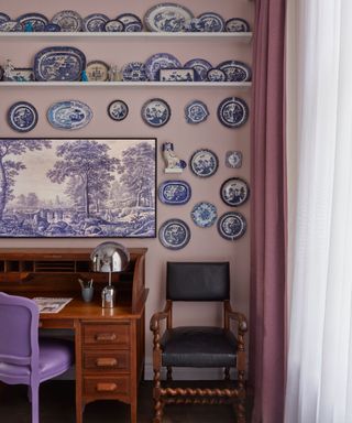 lilac pink hotel bedroom with a large collection of blue and white porcelain wall plates and a frame tv hung above a wooden vintage desk with a lilac chair and modern silver desk lamp