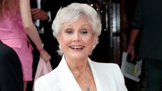 Portrait of Angela Rippon smiling wearing a white blazer with grey short hair, smokey eye and a rosy pink lip