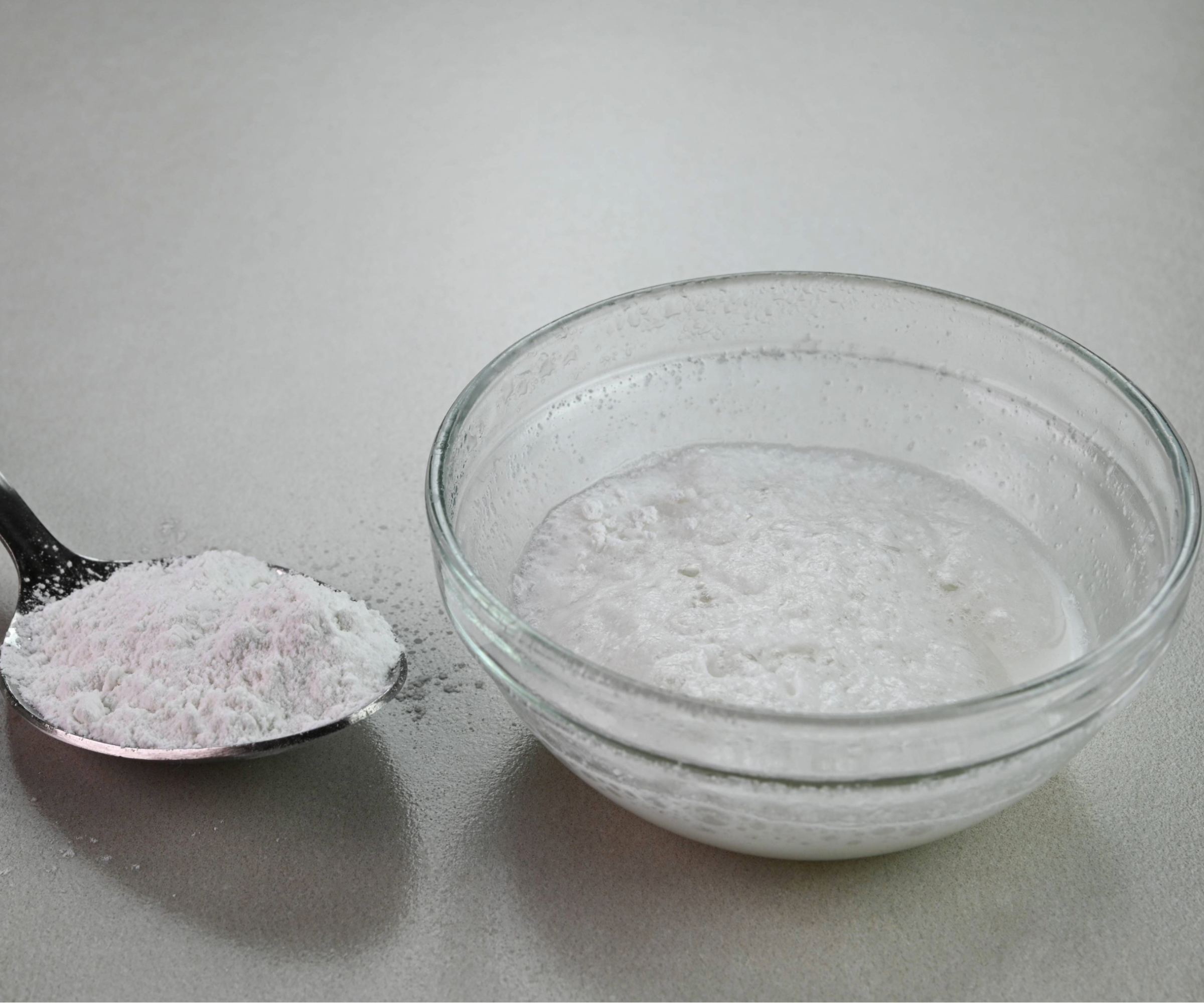 A spoon of baking powder next to a bowl of vinegar and baking soda
