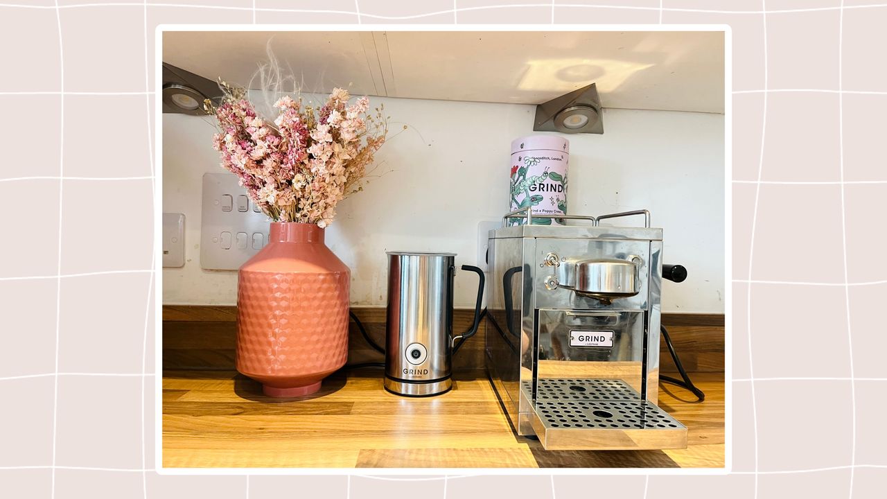 Grind One Pod coffee maker and milk frother on a wooden kitchen counter besides a pink vase of dried flowers.