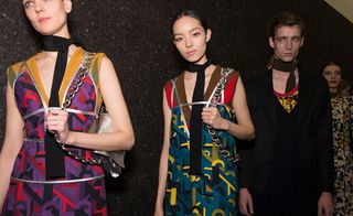 Four models wearing looks from Prada's collection. One female model is wearing a black neck scarf, orange and purple patterned dress with silver detail and she is carrying a silver bag with chain handle. Another female model is wearing a black neck scarf, brown and teal patterned dress with silver detail and she is carrying a bag with a chain handle. The third model is male and he is wearing a dark coloured neck scarf, yellow, red and black top and black suit jacket. And the fourth model is female and she is wearing a a white patterned piece with gold detail