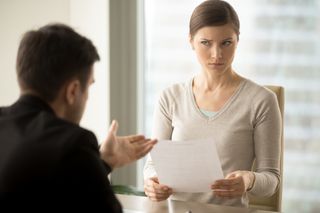 A woman holding a piece of paper with a skeptical look on her face as she listens to a man speaking to her from the other side of the table.