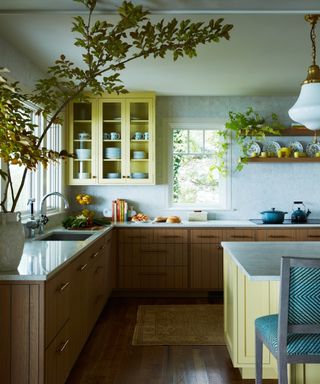 yellow and marble kitchen with wooden cabinets, pops of blue, hardwood floor, rug, marble countertops, blue upholstered bar stool, floating shelf with yellow cups and saucers, patterned plates, yellow wall cabinet,