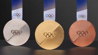 A set of the gold, silver, and bronze medals for the Milan 2026 Winter Olympics, featuring a distinctive circular design with a recessed, angled geometric cut across the center and the Olympic rings.