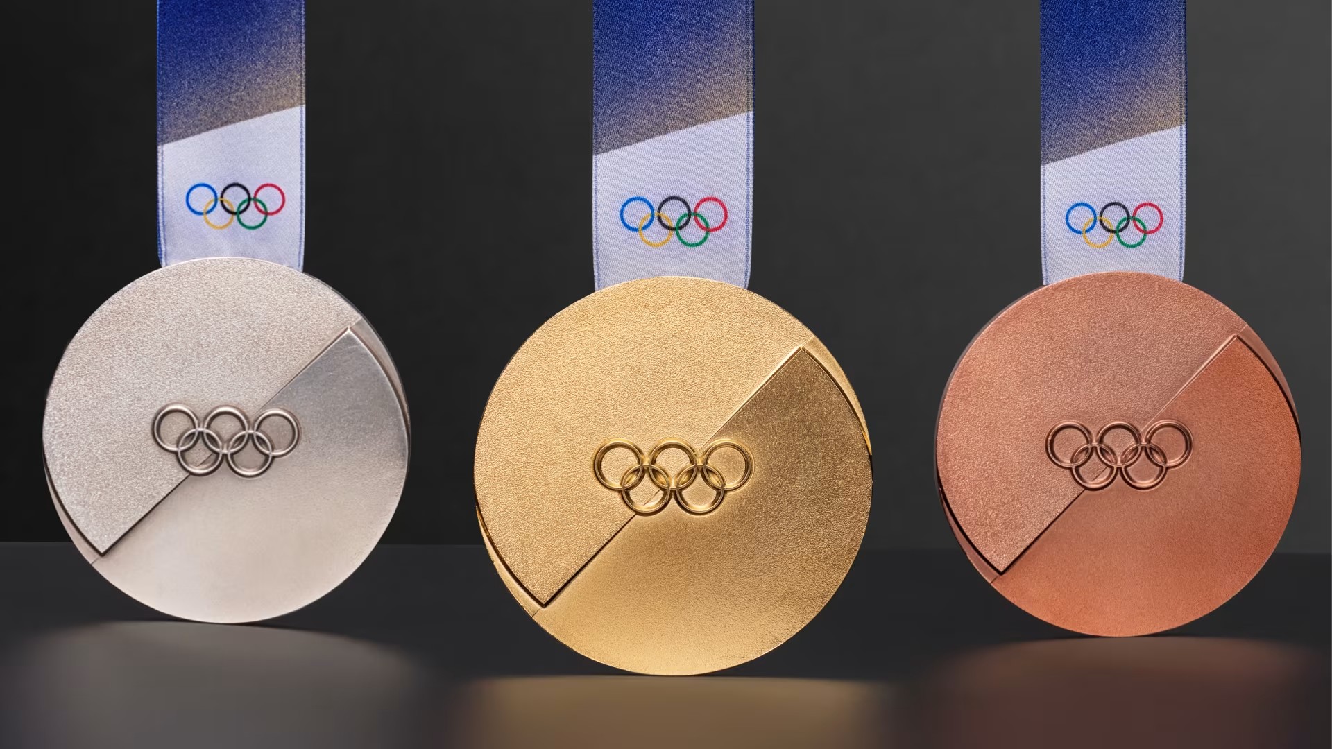 A set of the gold, silver, and bronze medals for the Milan 2026 Winter Olympics, featuring a distinctive circular design with a recessed, angled geometric cut across the center and the Olympic rings.