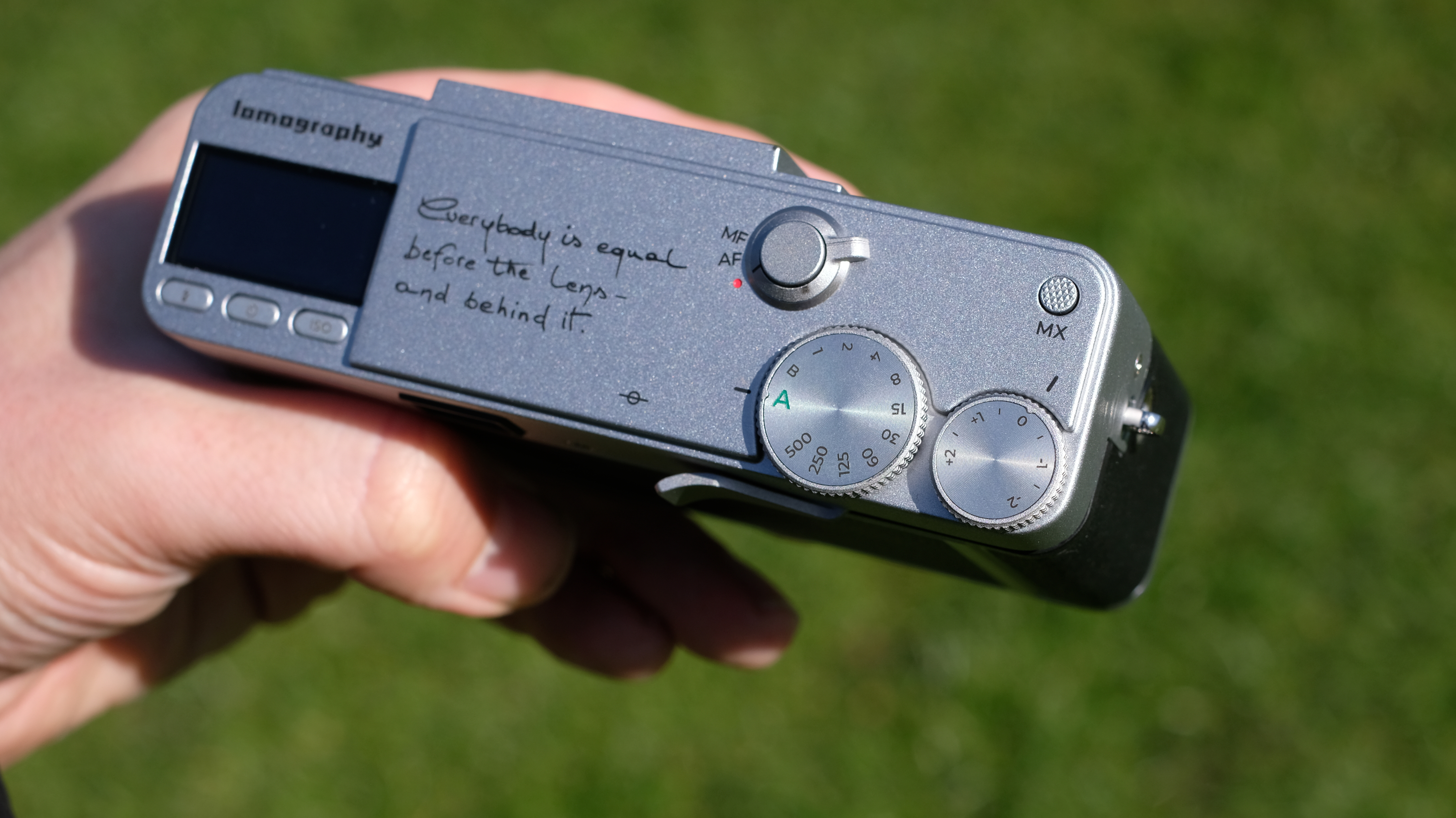 Photograph of the Lomography Lomo MC-A, with grass visible in background, showing top plate dials, engraving and screen