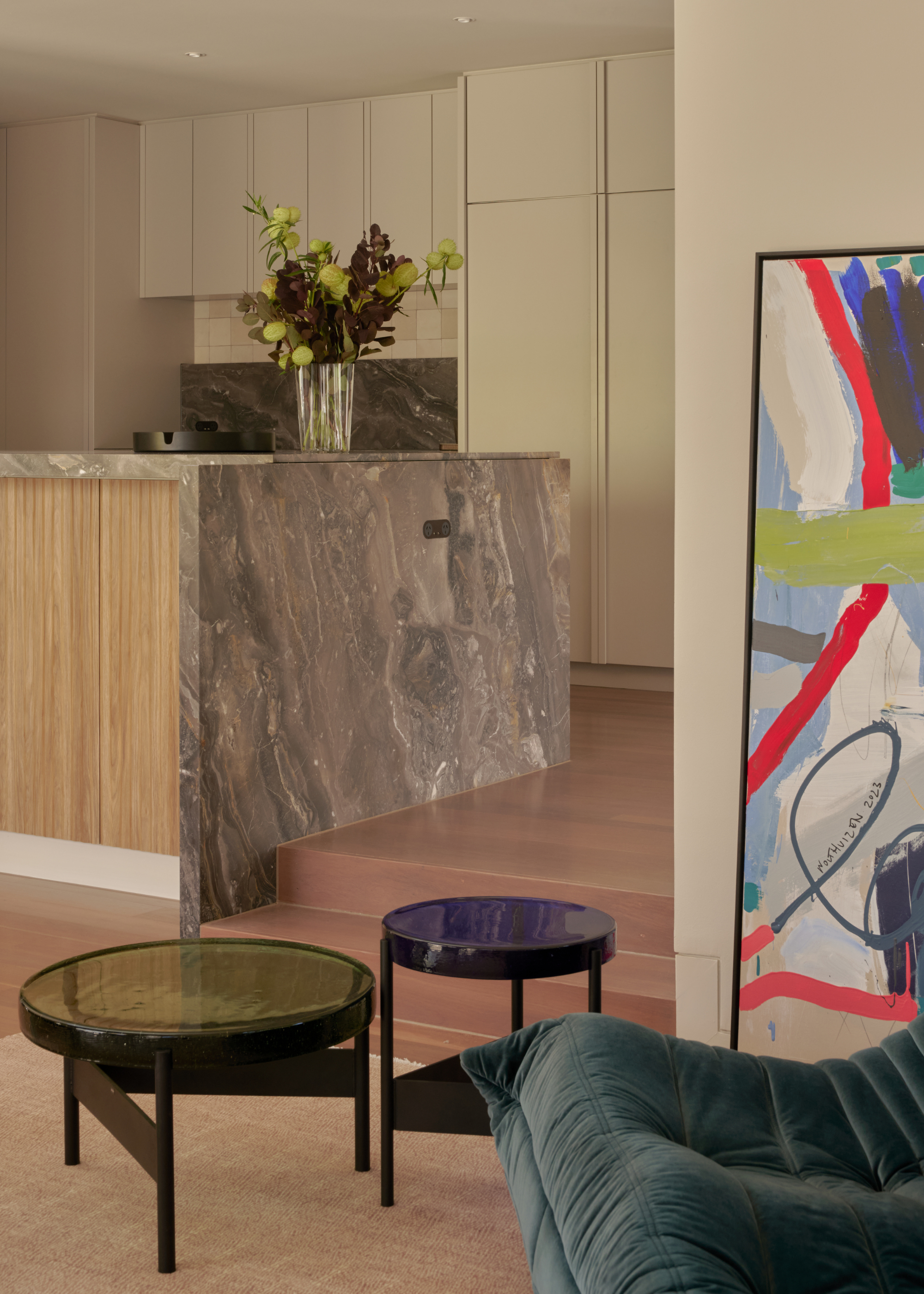 A kitchen with a stone island, a vase of flowers and some wood steps leading down to an open living room with a pair of glass coffee tables, a blue togo, and a leaning painting