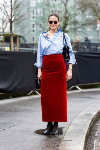 Eliza Huber wears black tights, black leather pumps, black sunglasses, a black leather bag, a light blue shirt and a high waist red suede long skirt outside Gucci show during the Milan Fashion Week Womenswear Fall/Winter 2025/2026 on February 25, 2025 in Milan, Italy.