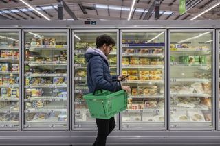 A man shopping in a supermarket whilst looking at his phone walking down the frozen aisle in front of a row of freezers.