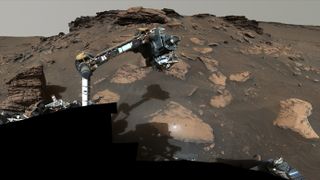 mars rover perseverance studies a rock with its robotic arm, with a rocky outcrop in the distance