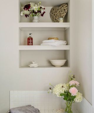 Alcove shelves in the bathroom with towels, flowers, soaps and a white dish above a white bath.