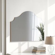 Image of an arched mirror on a white wall above a white marble counter. There is a stack of books and a glass lamp on the counter.
